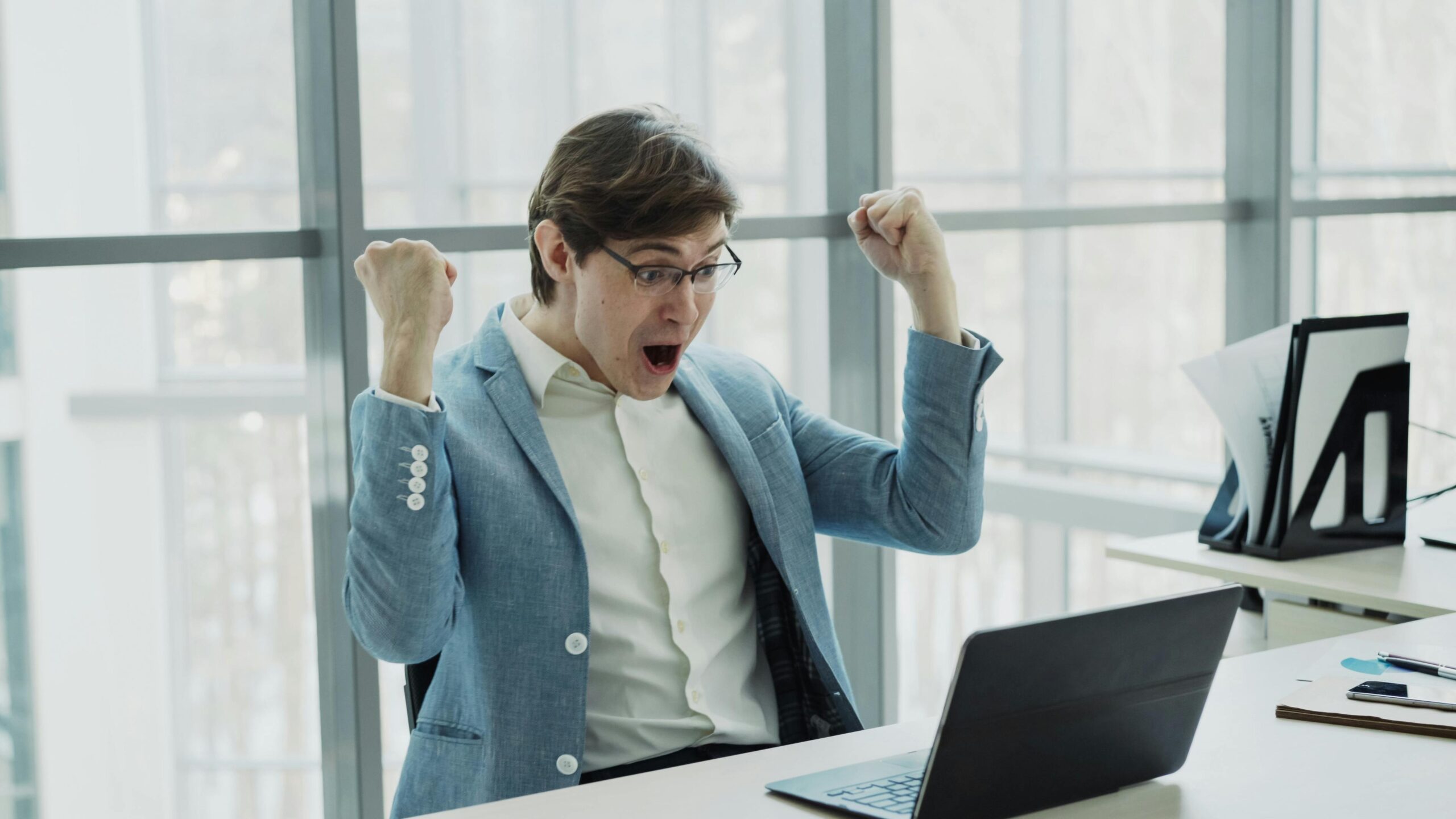 Young businessman in blue suit celebrates success at laptop indoors.